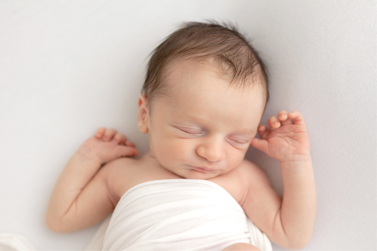Newborn baby boy sleeping on his back with hands up. Wrapped in a white swaddle in a white studio.