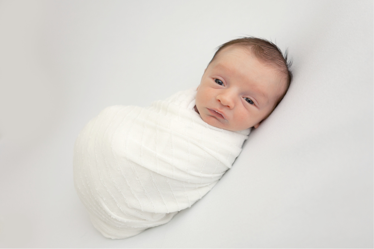 Newborn baby boy swaddled in a white swaddle. Laying on a white blanket with his eyes open.