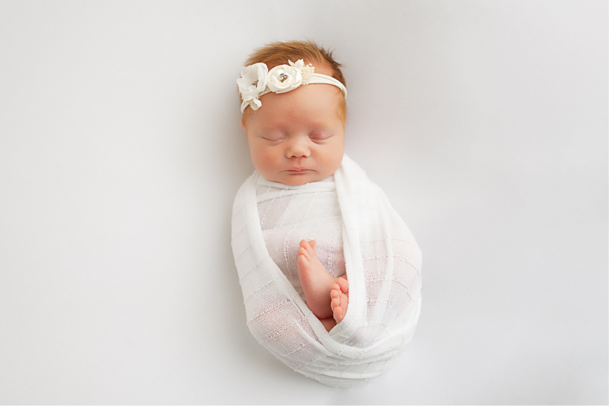 Sleeping red head newborn baby girl in studio newborn session. Wrapped in a white swaddle with white flower headband.