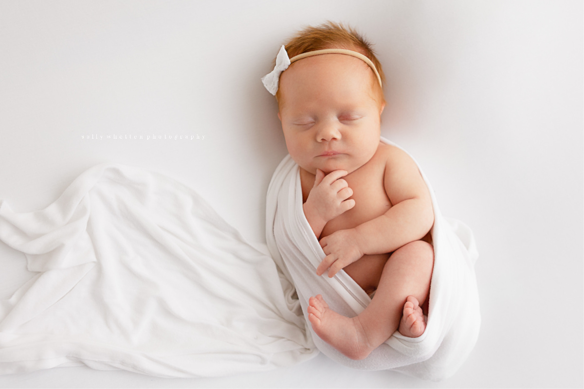 Baby girl with red hair wearing simple white hair bow. Sleeping in white swaddle on a white blanket.
