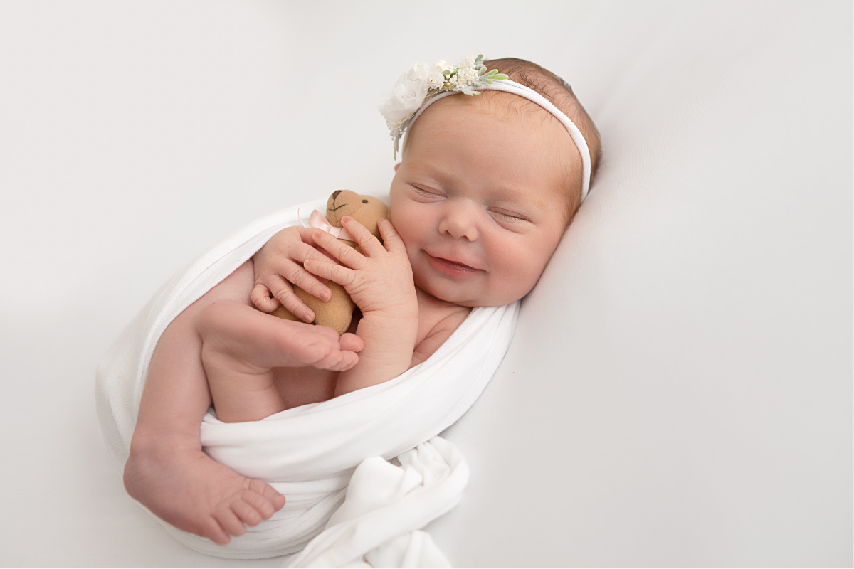 Newborn baby girl smiling while holding a tiny teddy bear. Baby girl is laying on a white blanket wrapped in a white swaddle.