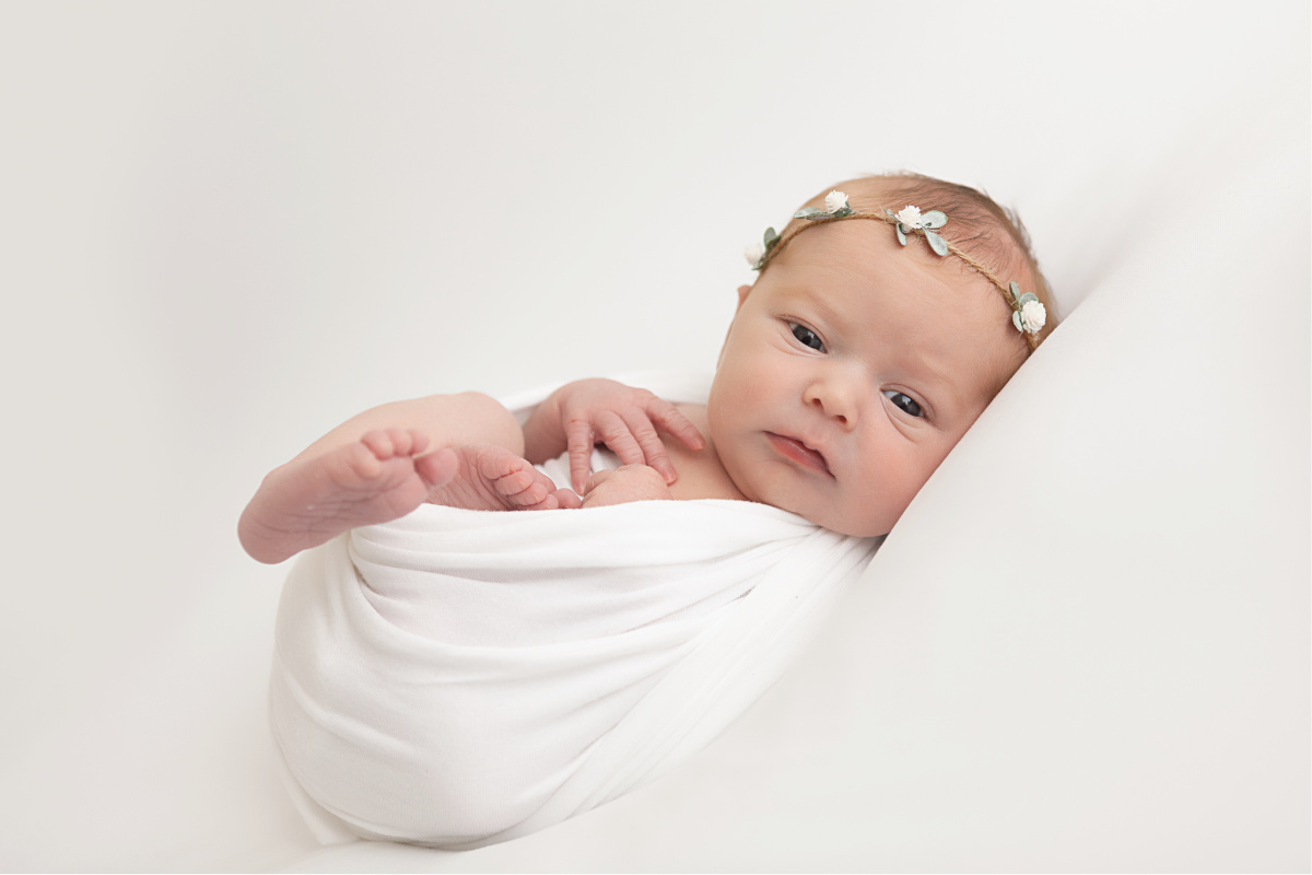 Newborn baby girl laying in white blanket. Wearing white flower and leaf headband. Awake baby photographed by Sally Whetten.