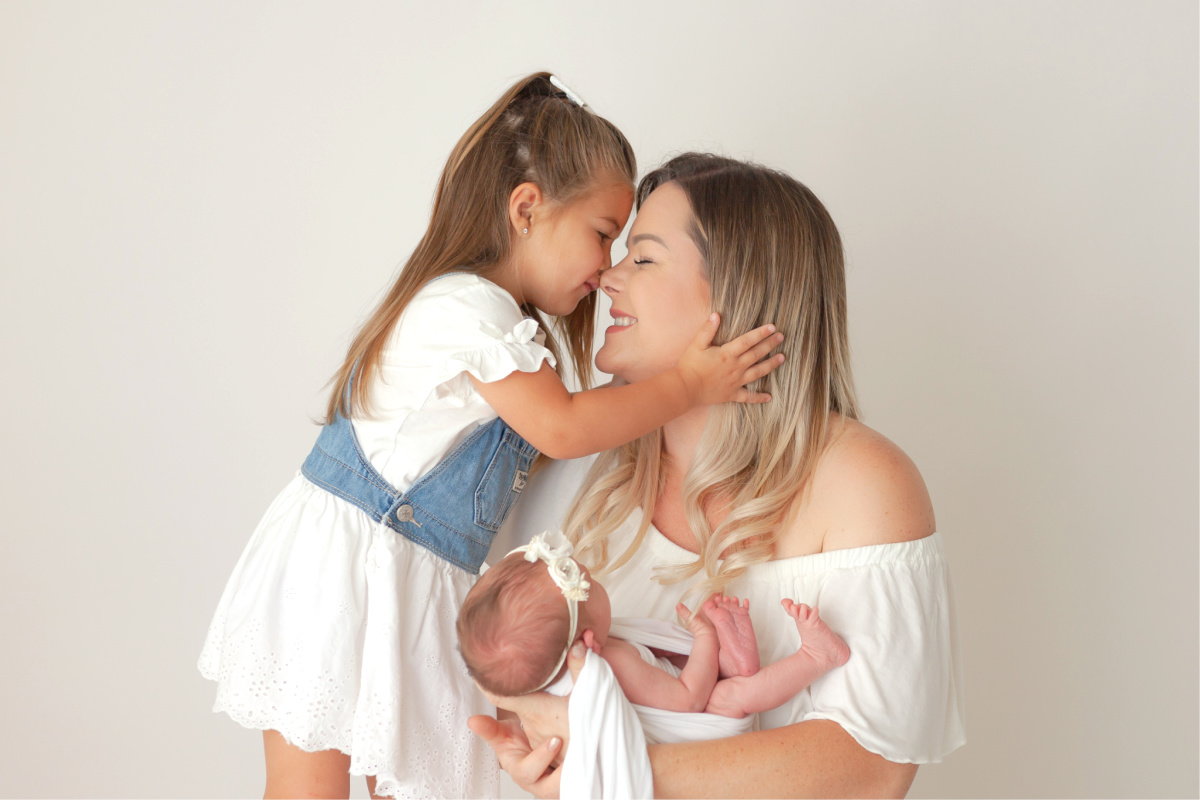 Mother getting a kiss from her older daughter. Mom snuggling new baby girl while getting hug from older sister. In a white studio.