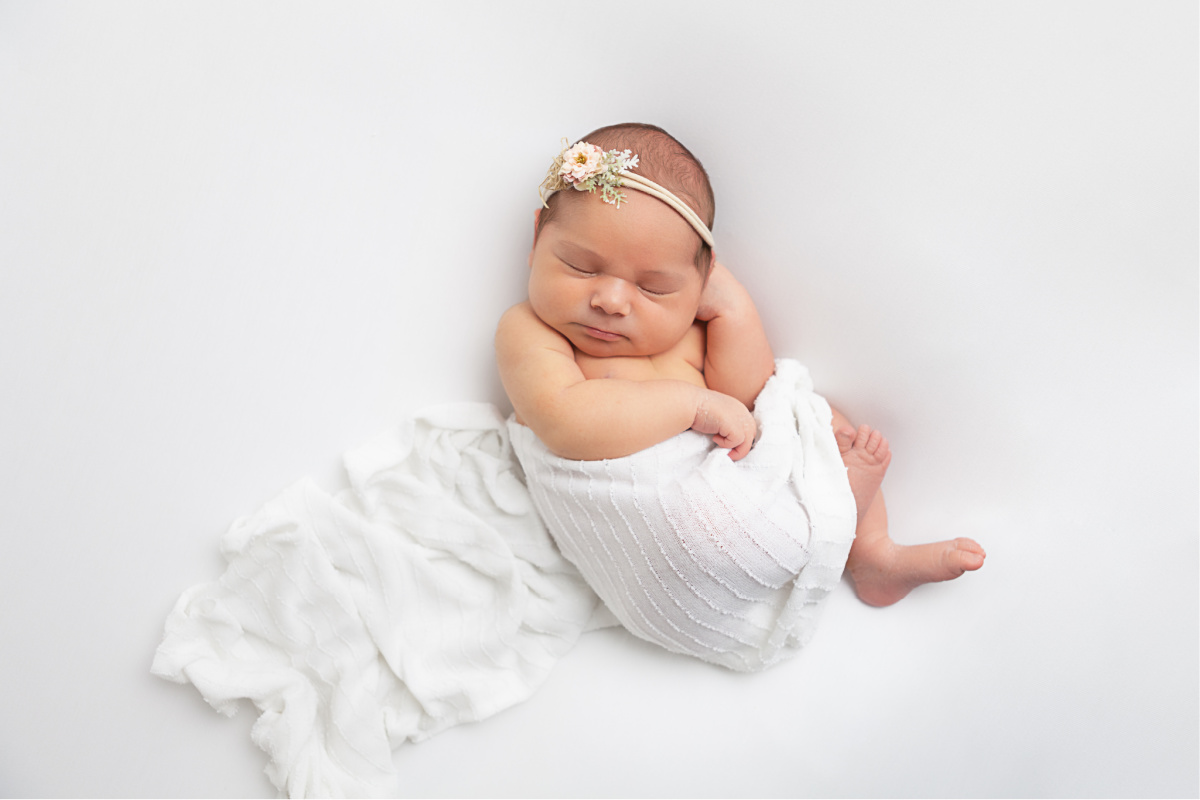 newborn girl laying on back on white backdrop, wrapped in white swaddle wearing a floral headband