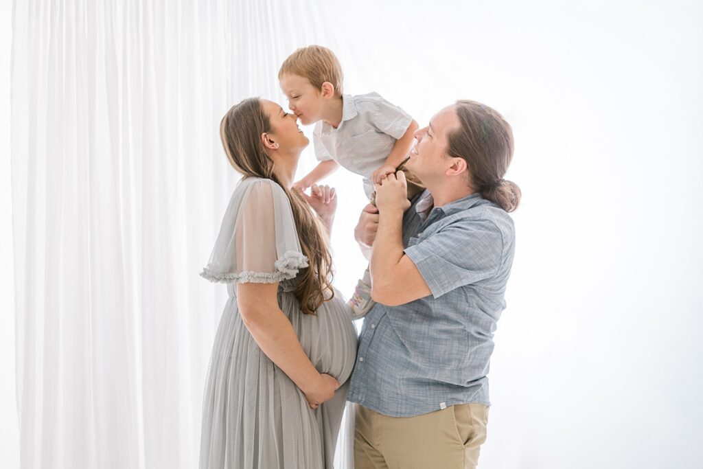 Dad holding toddler son while mom and son are nose to nose. Mom has her hand cradling her pregnant belly in beautiful white studio.