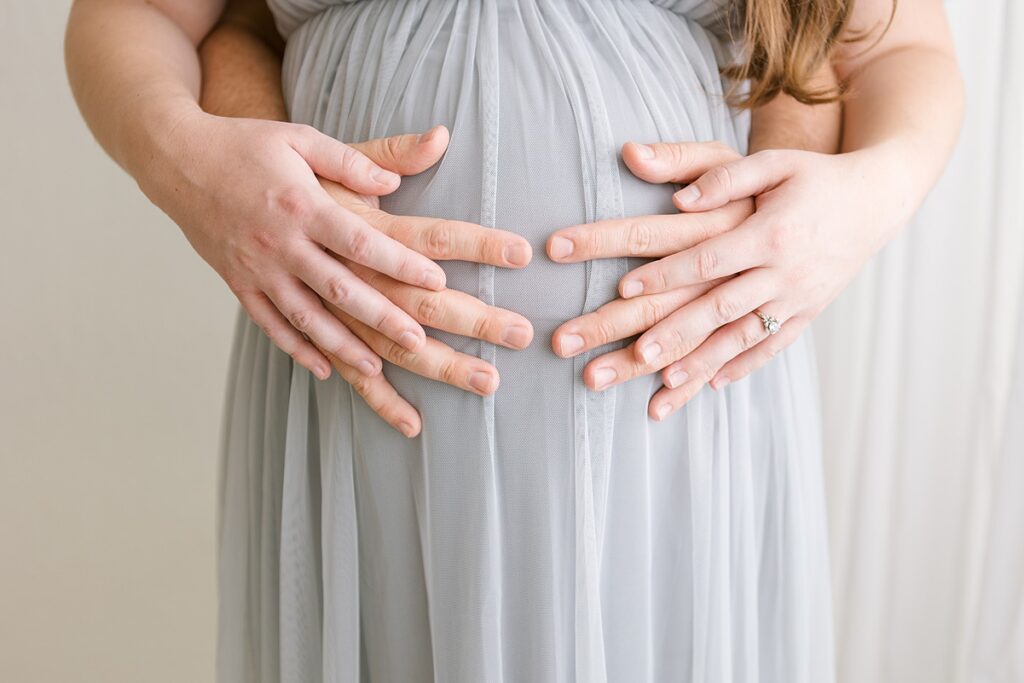 Dad standing behind mom and has his arms wrapped around her so all four of their hands are on her pregnant belly.