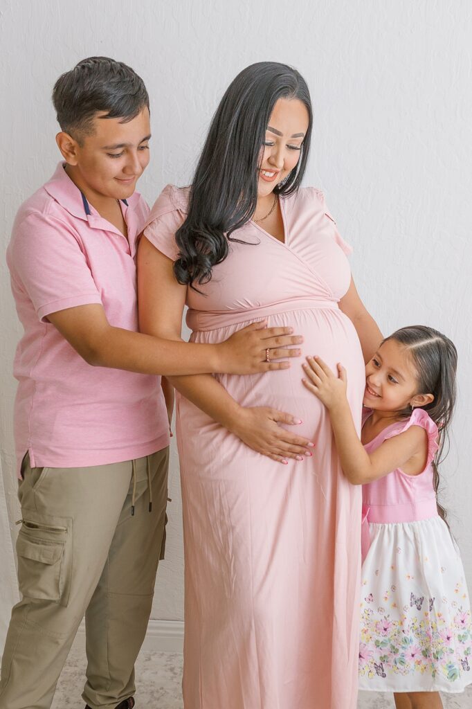 maternity photo of a mom in a pink dress with her older son and daughter next to her touching her belly