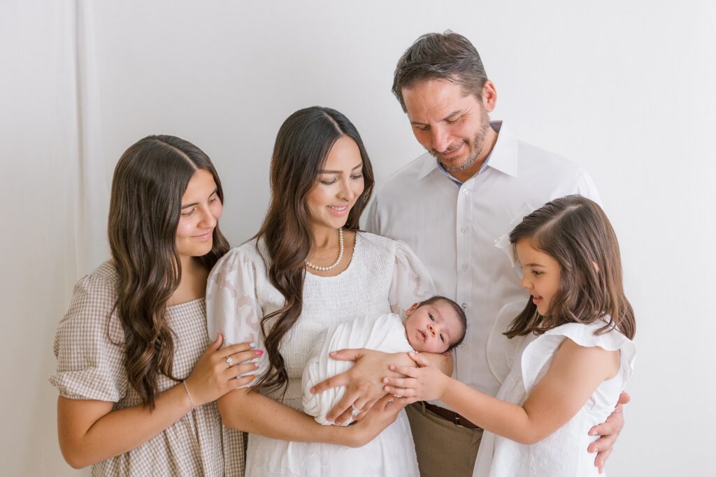 Family of 5 all looking at a newborn baby boy in a white studio portrait