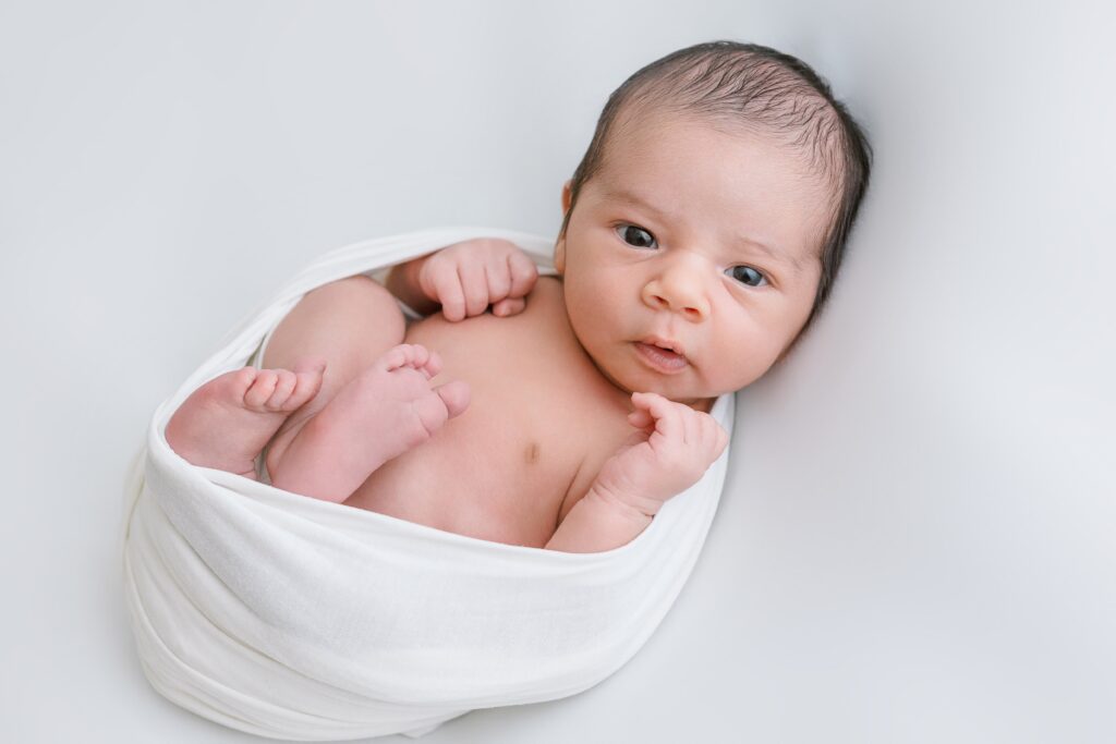 Awake newborn baby boy wrapped peacefully and laying on a white backdrop.