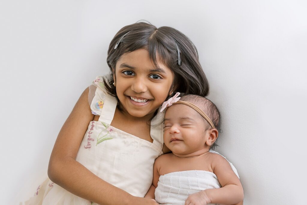 Big sister cuddling up to newborn sister on a white backdrop.