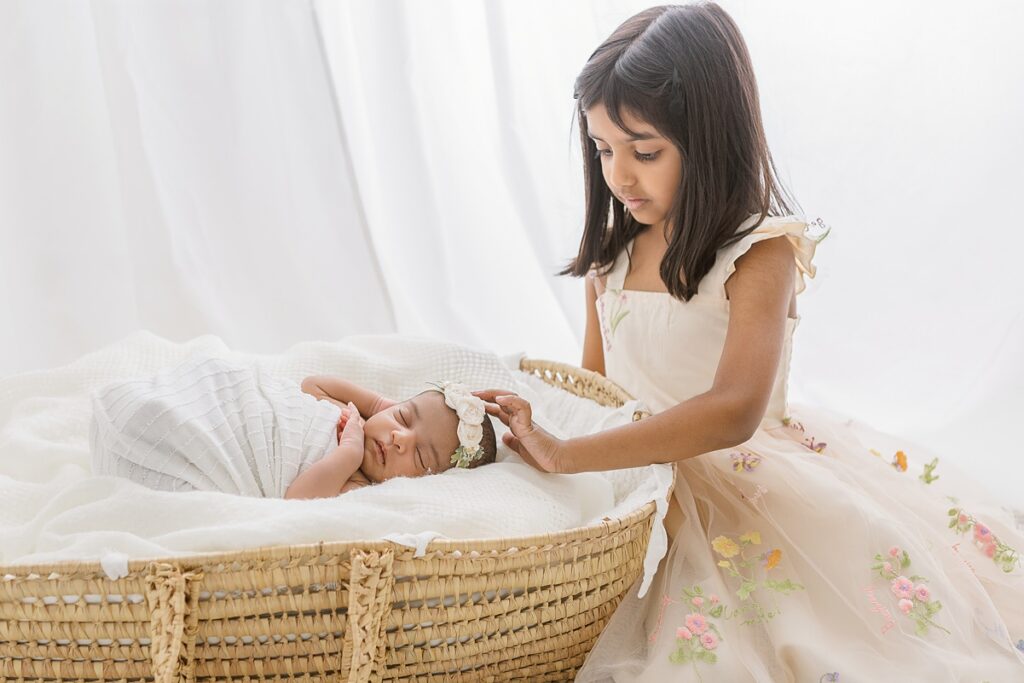 Big sister gently touching baby sister's head while she is laying in a moses basket in chandler newborn photography studio.