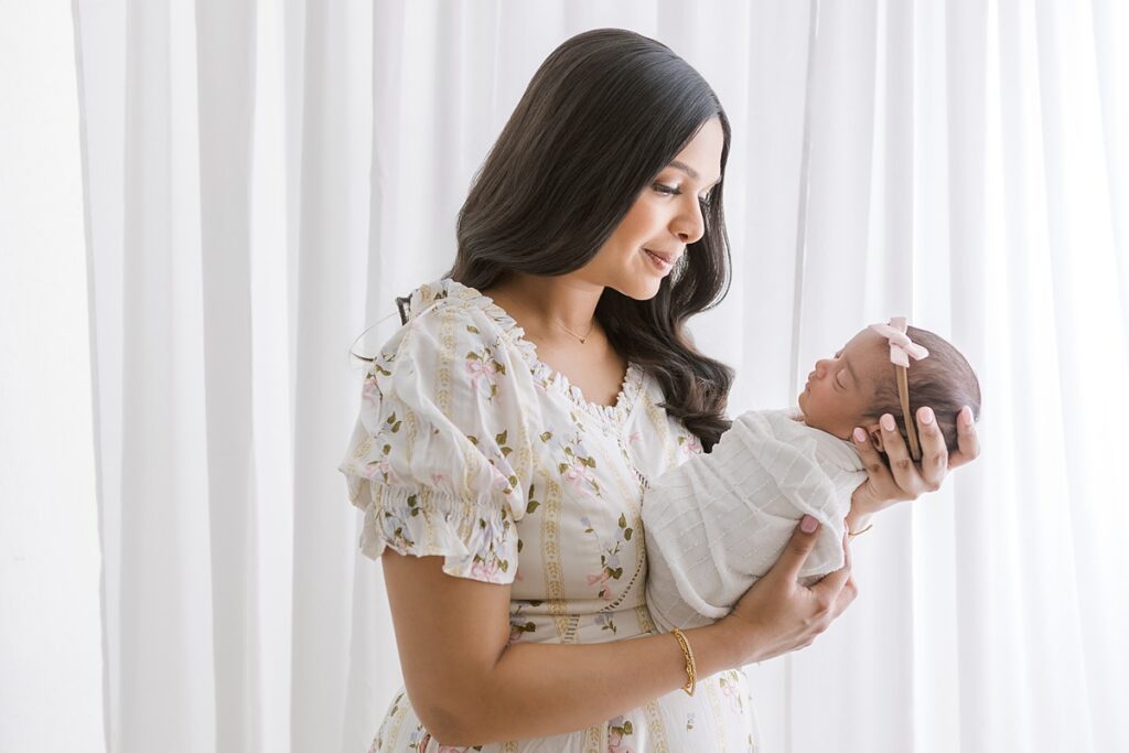 Side view of mom holding newborn daughter in front of a white curtain with soft glowing light.