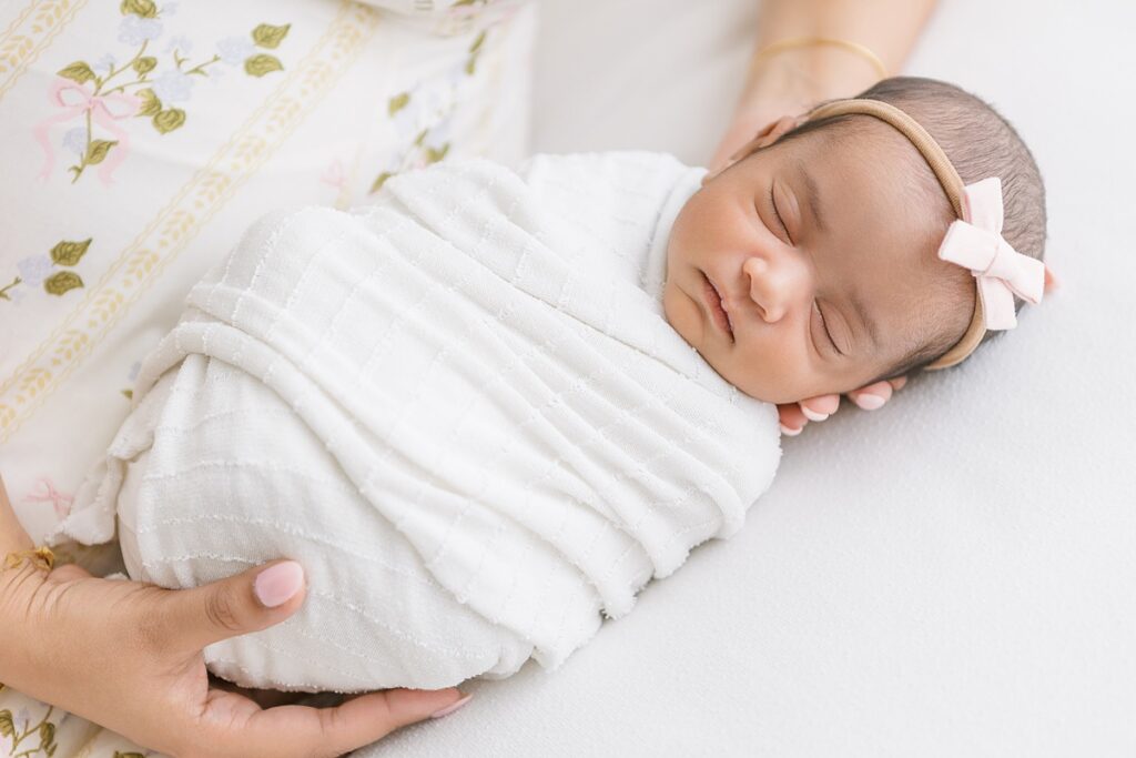 sweet newborn baby girl laying in her mothers hands wearing a white swaddle and white hair bow