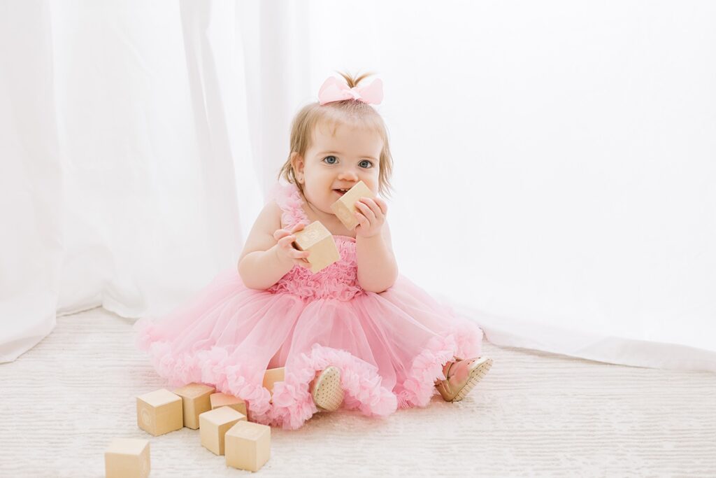 one year old little girl sitting in pink dress in front of a white curtain with soft glowing light, playing with wooden blocks for her mesa first birthday photo session