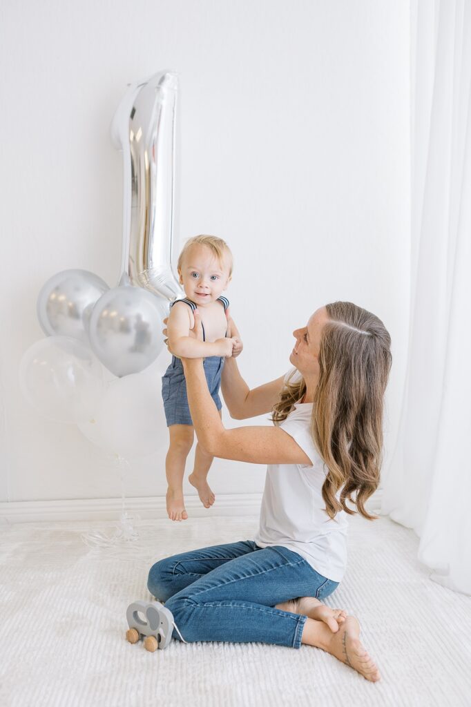 mom holding up little one year old in front of large silver number one balloon in white photo studio.