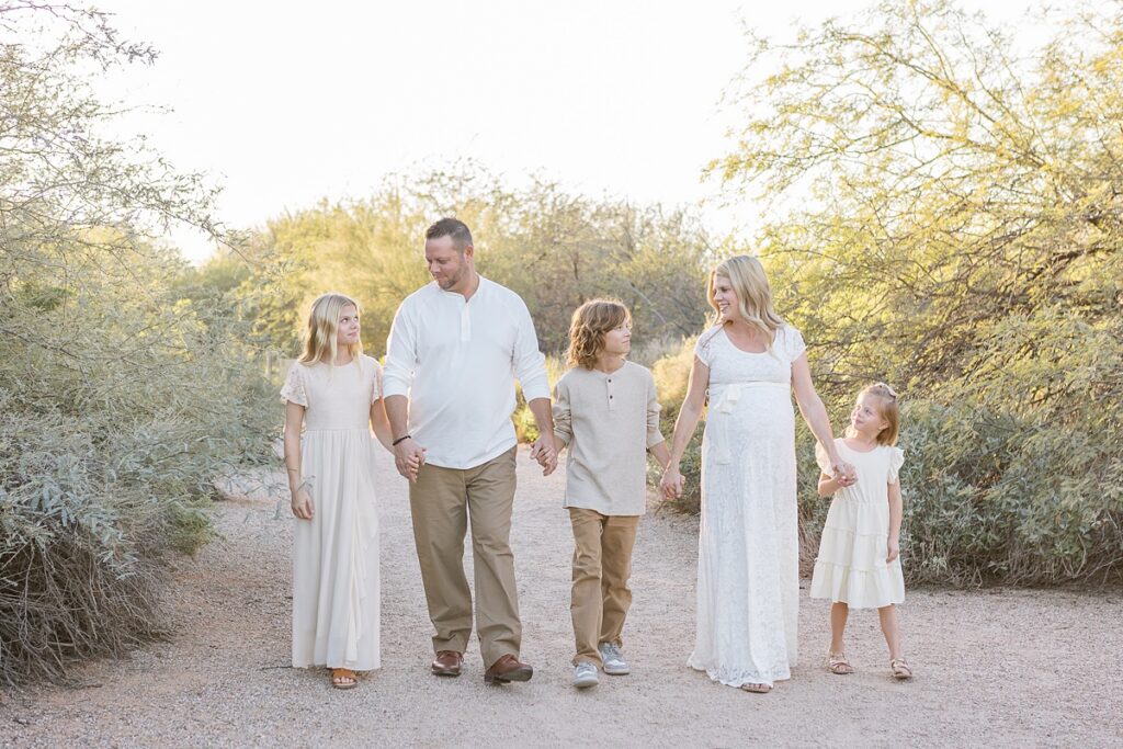Family of 5 in neutral clothes holding hands in a line and walking in beautiful desert in the evening.