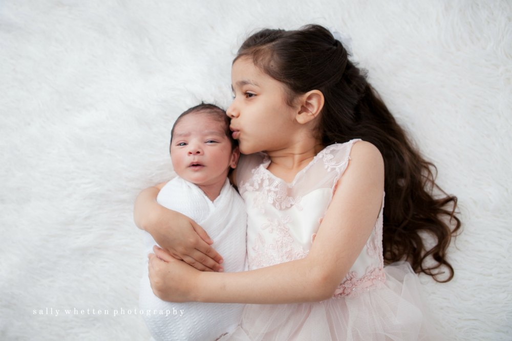big sister in pink dress kissing newborn brother on fur rug