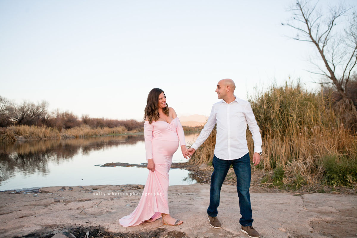 couple taking maternity photos by the river at sunset