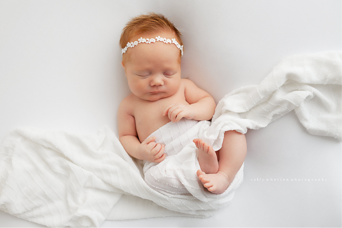 Minimal newborn portrait of a baby girl in white swaddle. She is on a white background wearing a simple white headband.