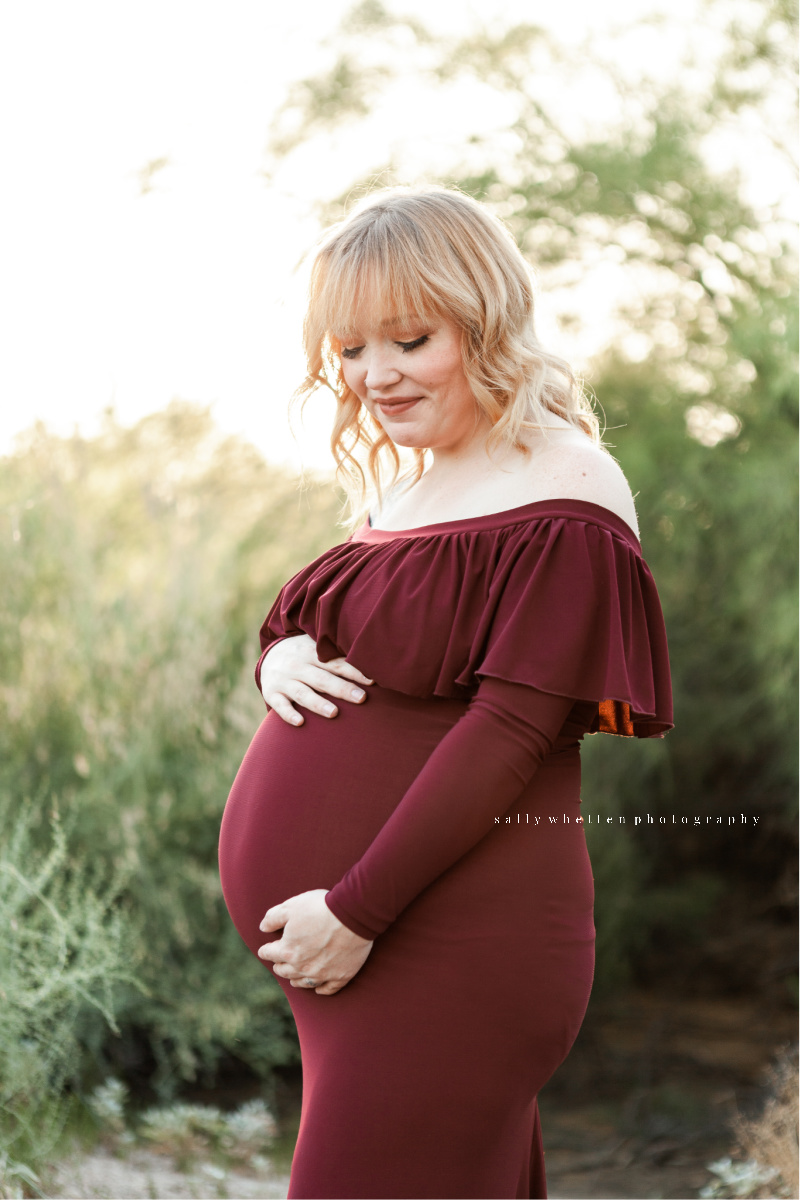Expecting mother looking at her belly. Wearing a maroon dress in a sunset photo session. Taken at the Gilbert Riparian Preserve.