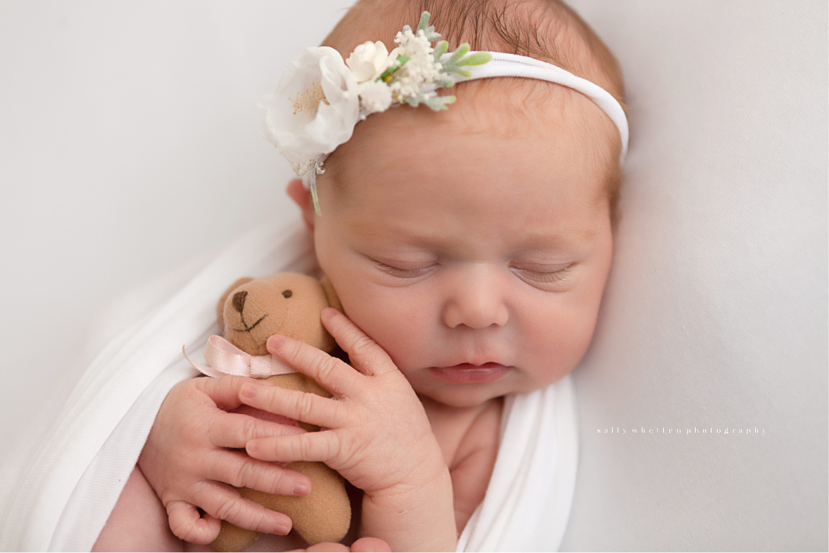 Newborn baby girl wearing a white flower headband. She is hugging a little teddy bear. Photographed on a white backdrop in a studio.
