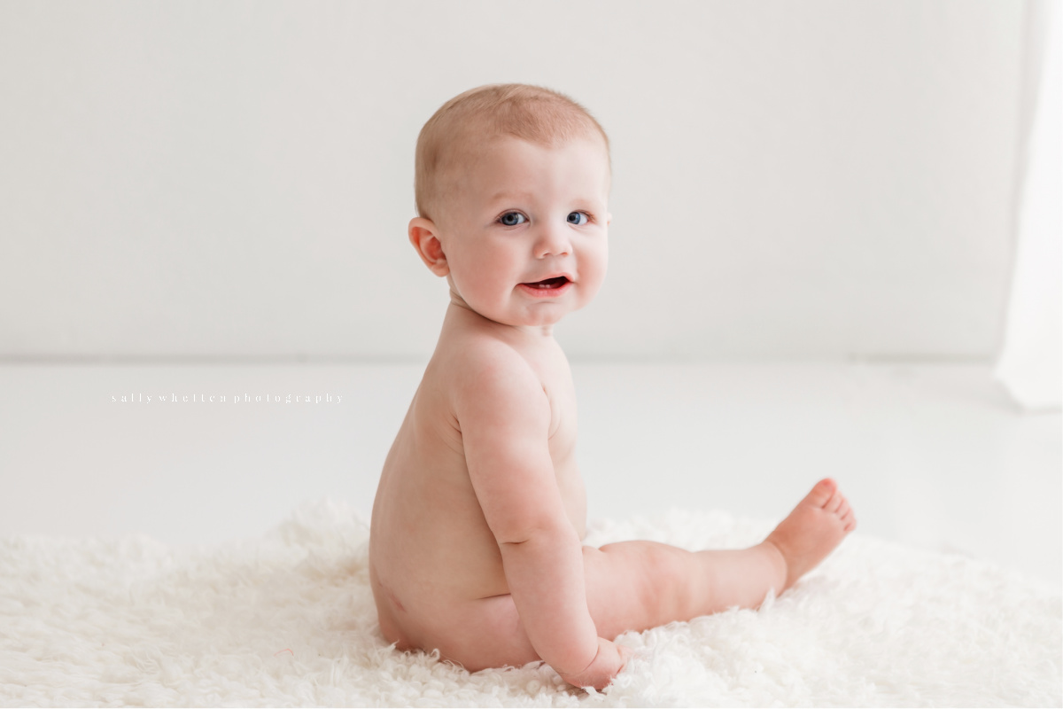 Six month old baby boy sitting by himself on a white fur rug. Looking over his shoulder and smiling.