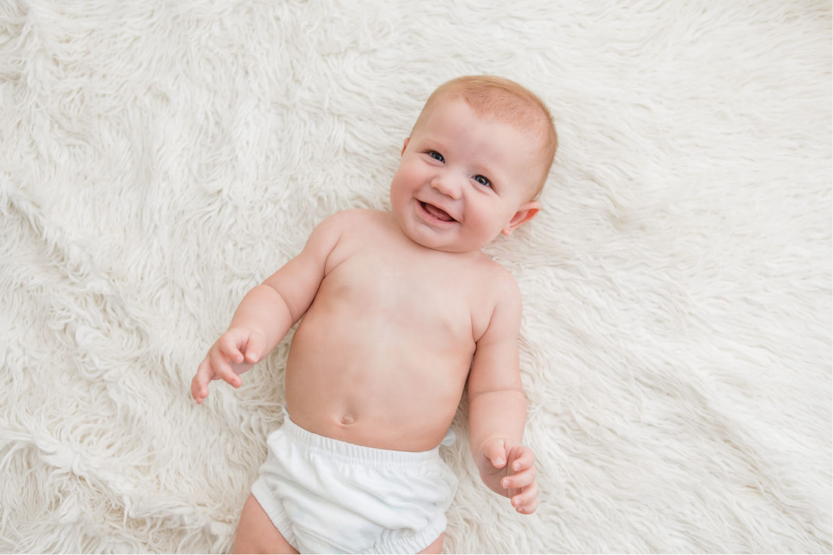 Six month old baby boy laying on white rug. Wearing a white diaper cover. Looking up at the camera and smiling.
