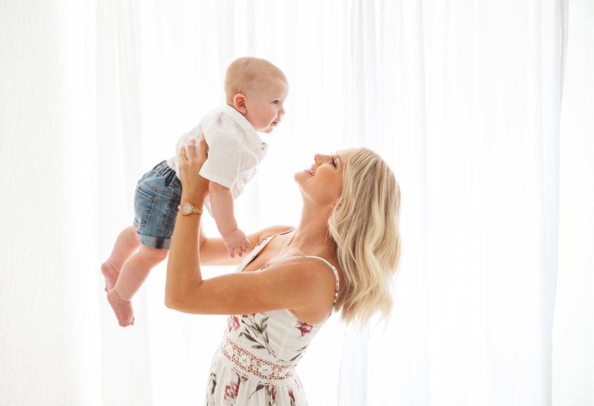 Mother holding six month old baby boy up and smiling at him. Photographed in white studio by photographer Sally Whetten Photography.