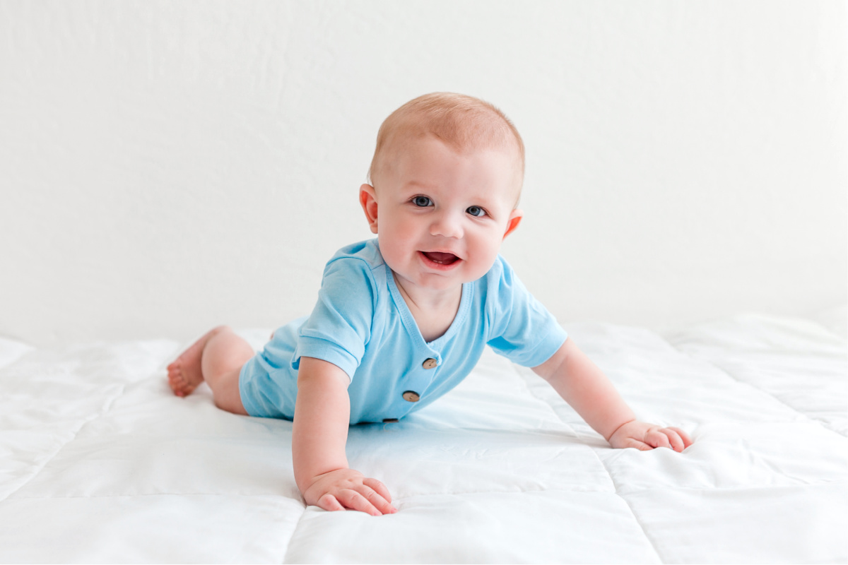 Six month old baby boy in a light blue outfit. On his tummy on a white blanket. Photographed in a white studio by Sally Whetten.