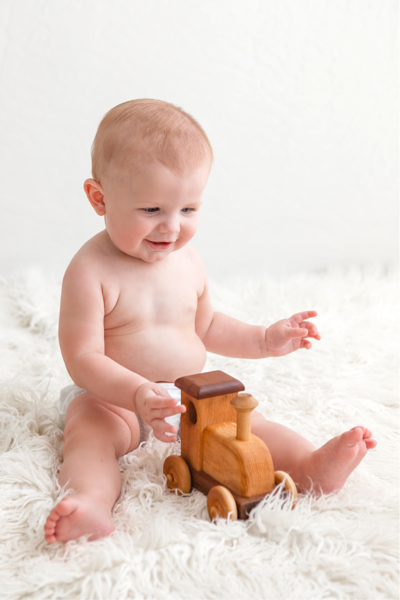 Six month old studio session where baby boy is sitting alone. He is playing with a natural wood train on a white fur blanket.