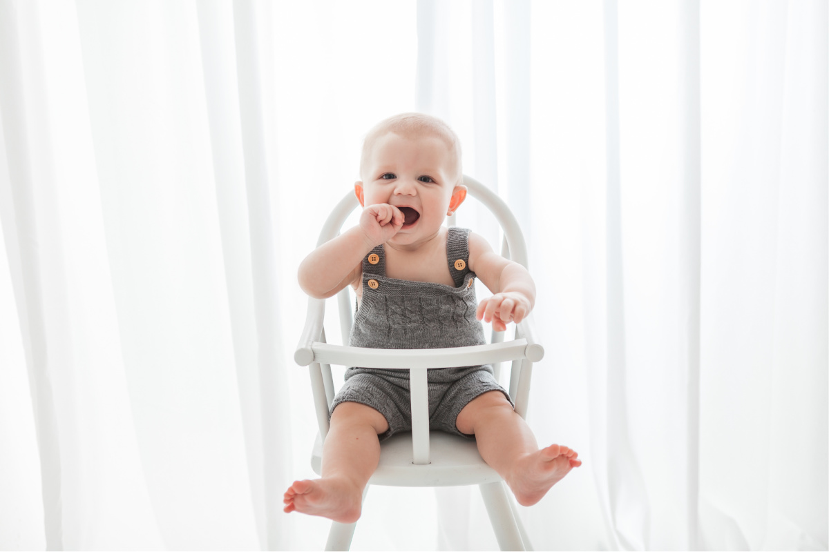 Six month old baby boy wearing gray knitted overalls. Sitting in a white high chair in front of a bright white curtain. In studio session by photographer Sally Whetten.