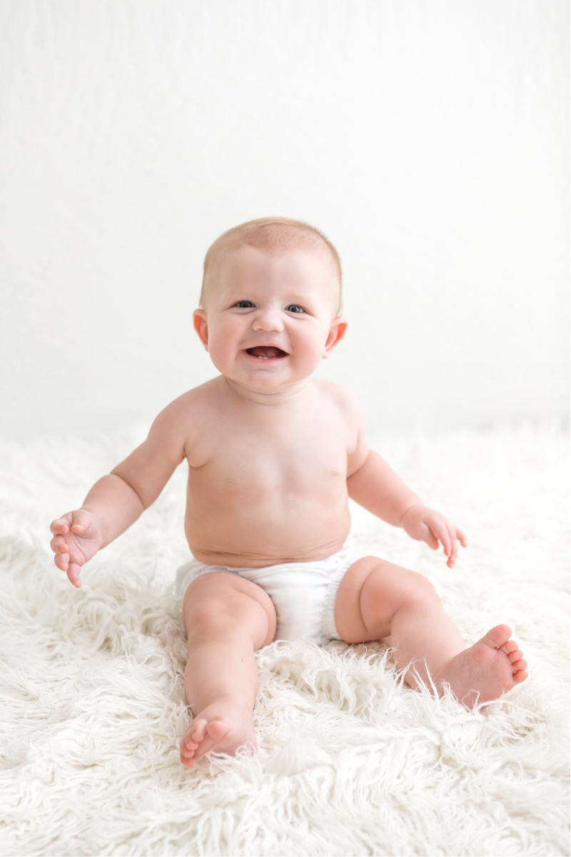 Six month old studio session photographed by Sally Whetten Photography. Six month old baby boy in white diaper cover. Sitting on a white fur rug and smiling.