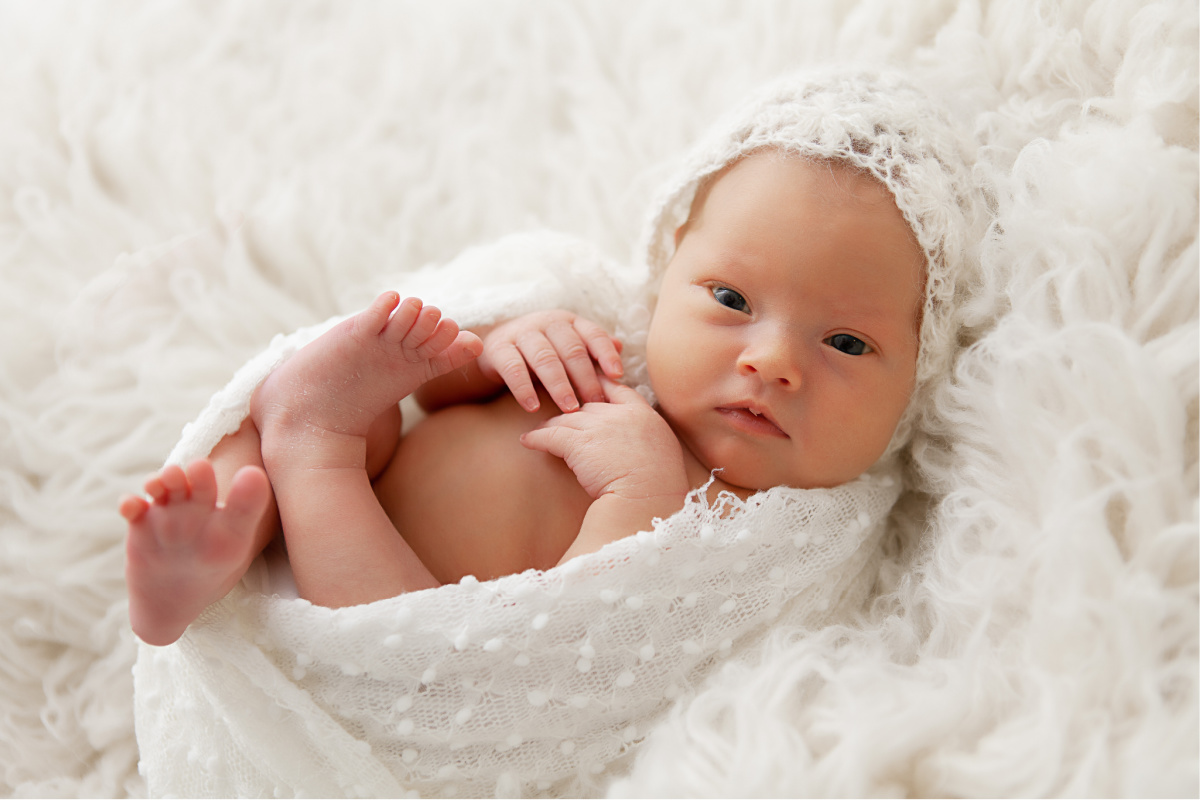 newborn baby girl wearing a bonnet awake lying in white fur wrapped with knit blanket