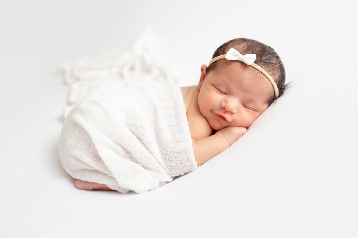 newborn baby girl sleeping on her tummy with white bow headband, smiling in her sleep