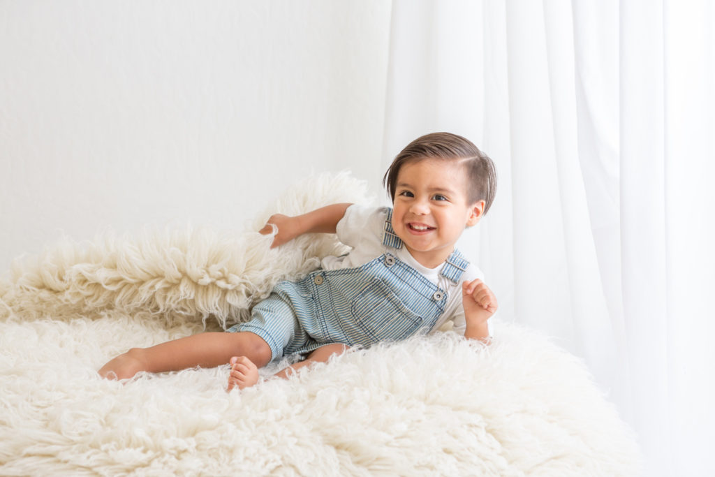 two year old boy laying on a white fur rug smiling
