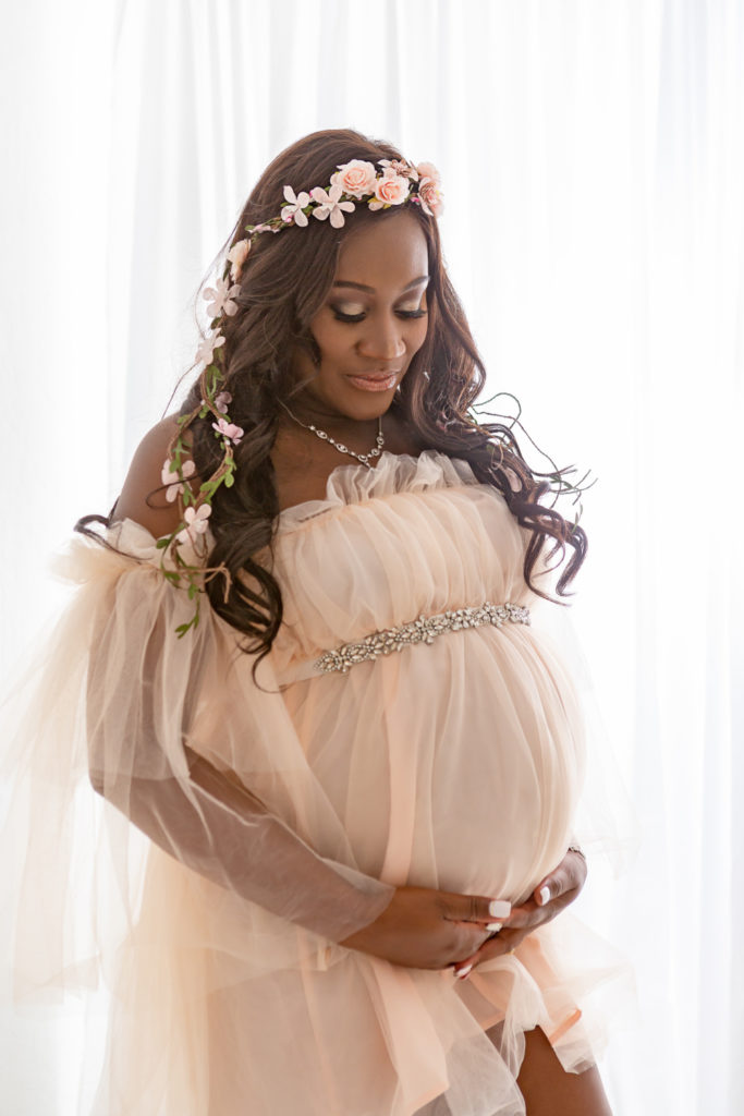 beautiful pregnant woman of color wearingn light blush dress wearing a flower crown. In a white photography studio.
