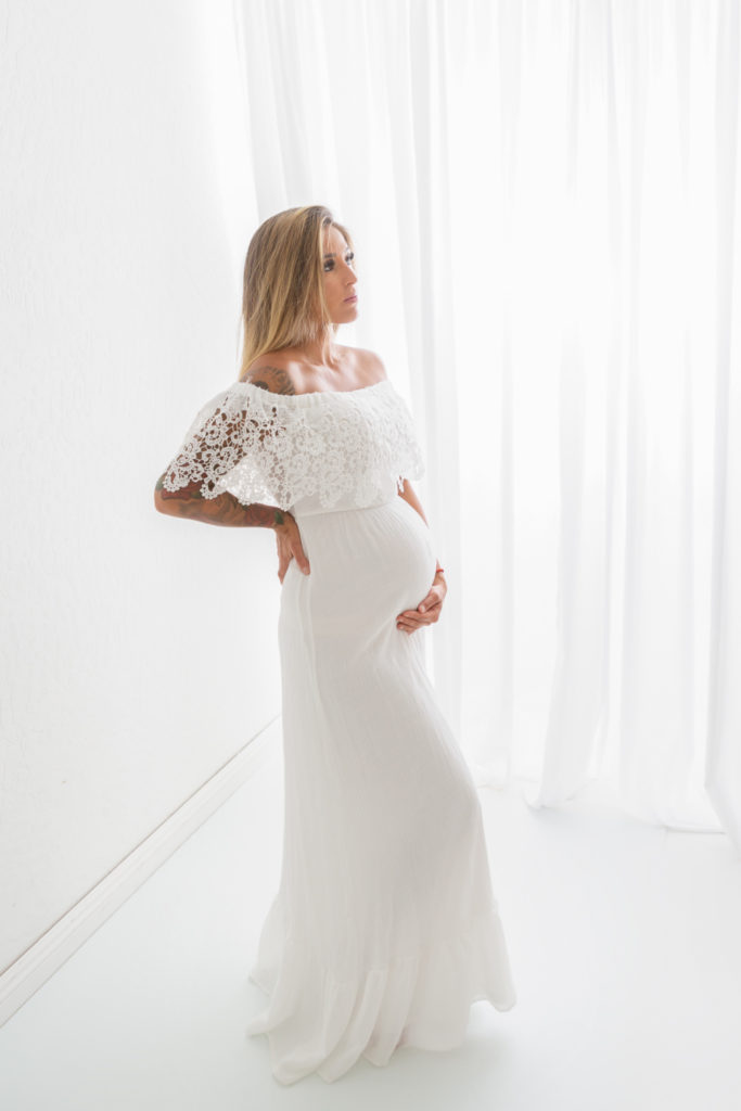 Pregnant woman in white dress standing in front of window with a white curtain. 