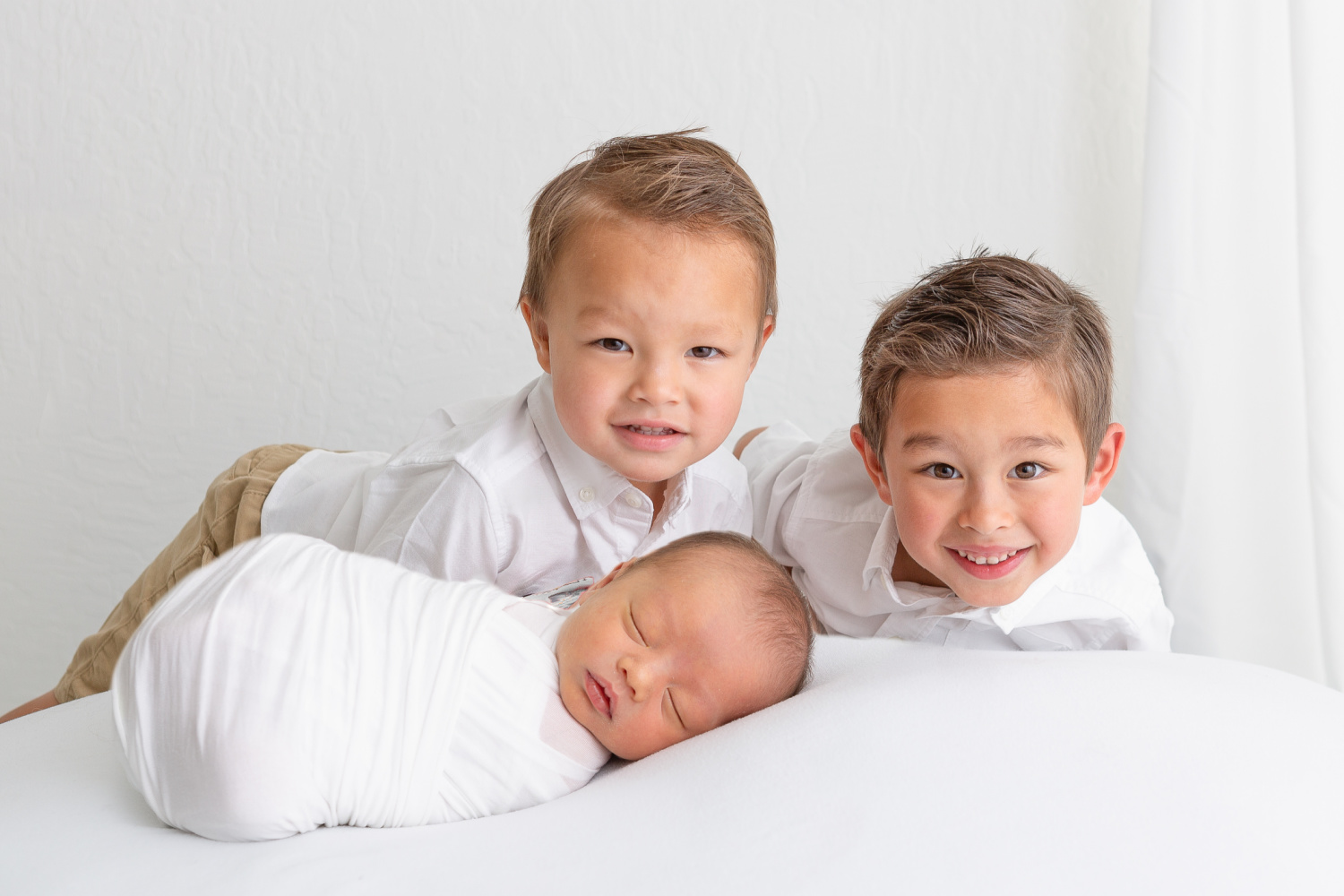 newborn baby boy wrapped in white with his two brothers behind him smiling