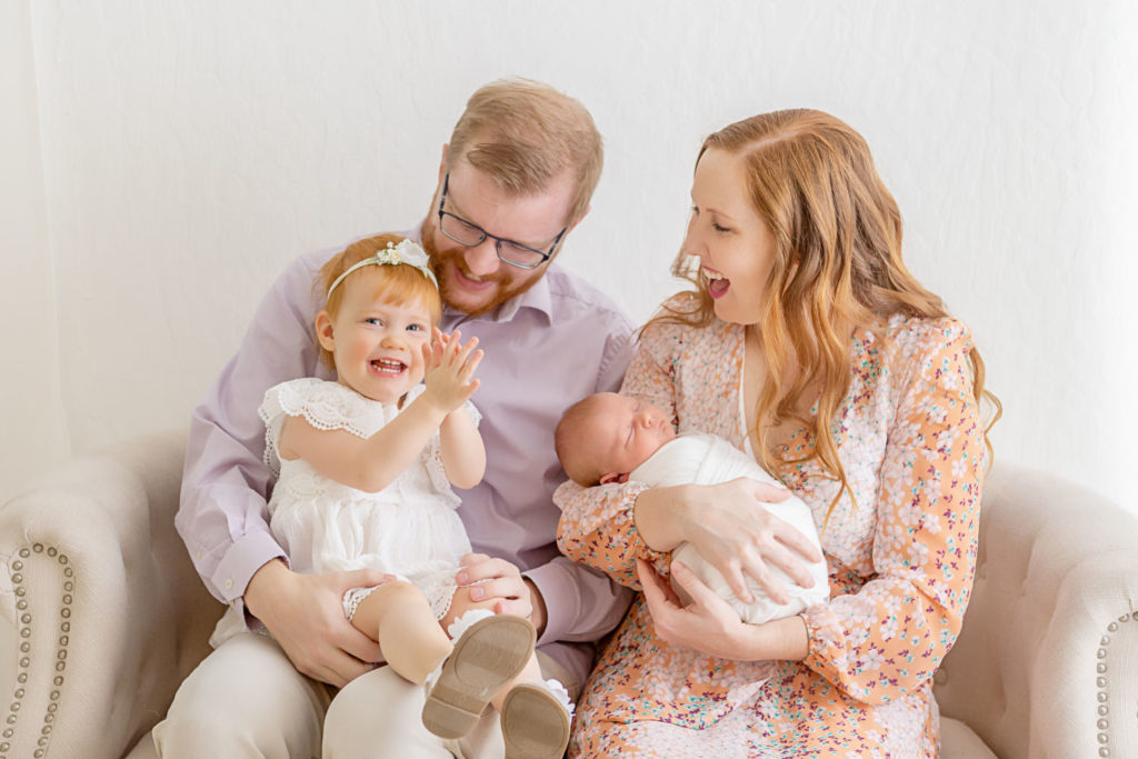 family of four in photo studio, sitting on a loveseat with newborn baby and happy toddler