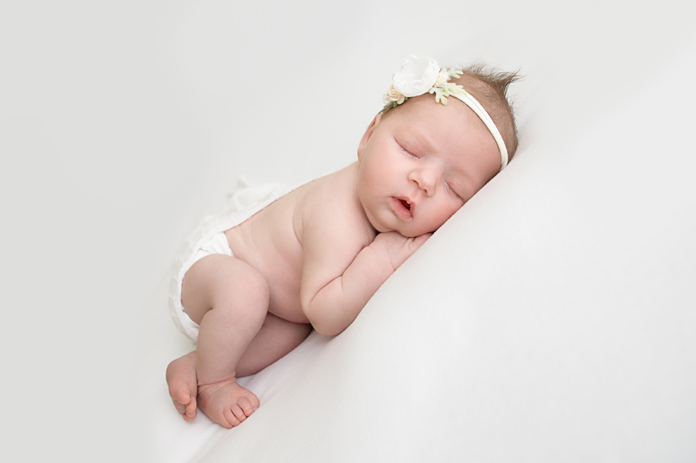 Sweet newborn baby girl asleep on her tummy. She is wearing a white floral headband and laying on a white backdrop in a Phoenix area newborn photography studio.