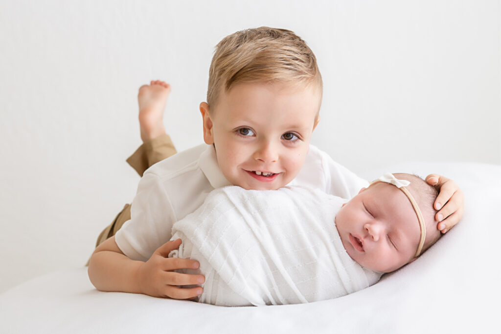 Newborn baby girl with her older brother cradling her in his hands. Both are laying on a white blanket in a Phoenix newborn photo studio.