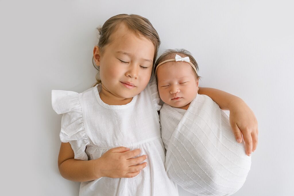 Big sister snuggles baby girl on a white bed in a bright, cozy studio.