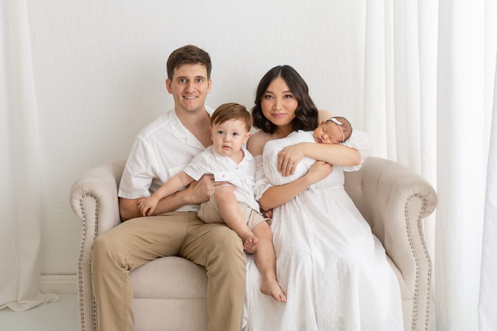 Phoenix baby photographer captures a joyful family moment in a white studio—mom, dad, toddler brother, and baby sister all snuggled together in a bright, loving setting.
