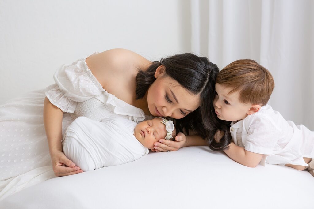 Moment of sweet connection between mom, her newborn, and toddler cuddled on a white blanket.