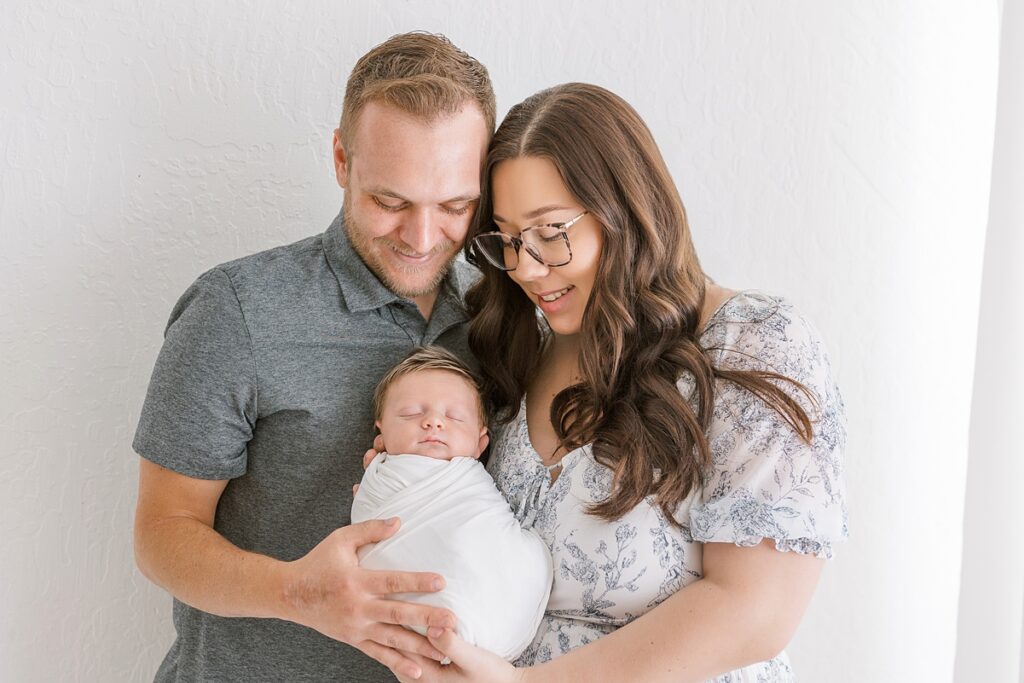 Parents holding their newborn baby boy and looking down at him with love in a soft, light-filled setting.