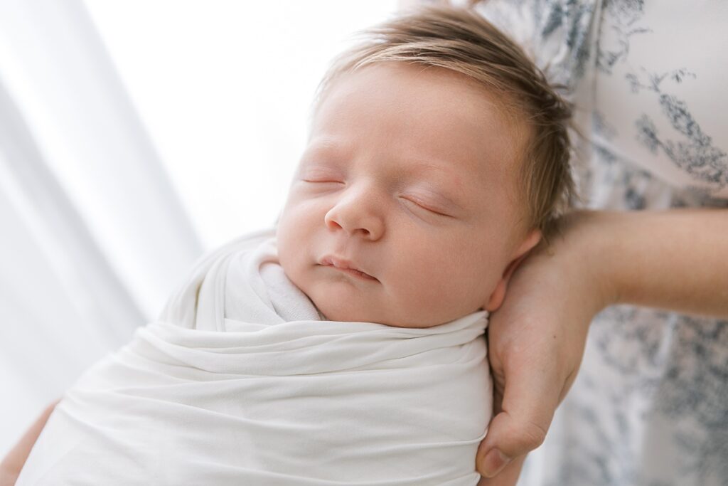 Newborn baby boy sleeping peacefully in his parents’ hands, surrounded by soft, beautiful light in Gilbert baby photographer studio.