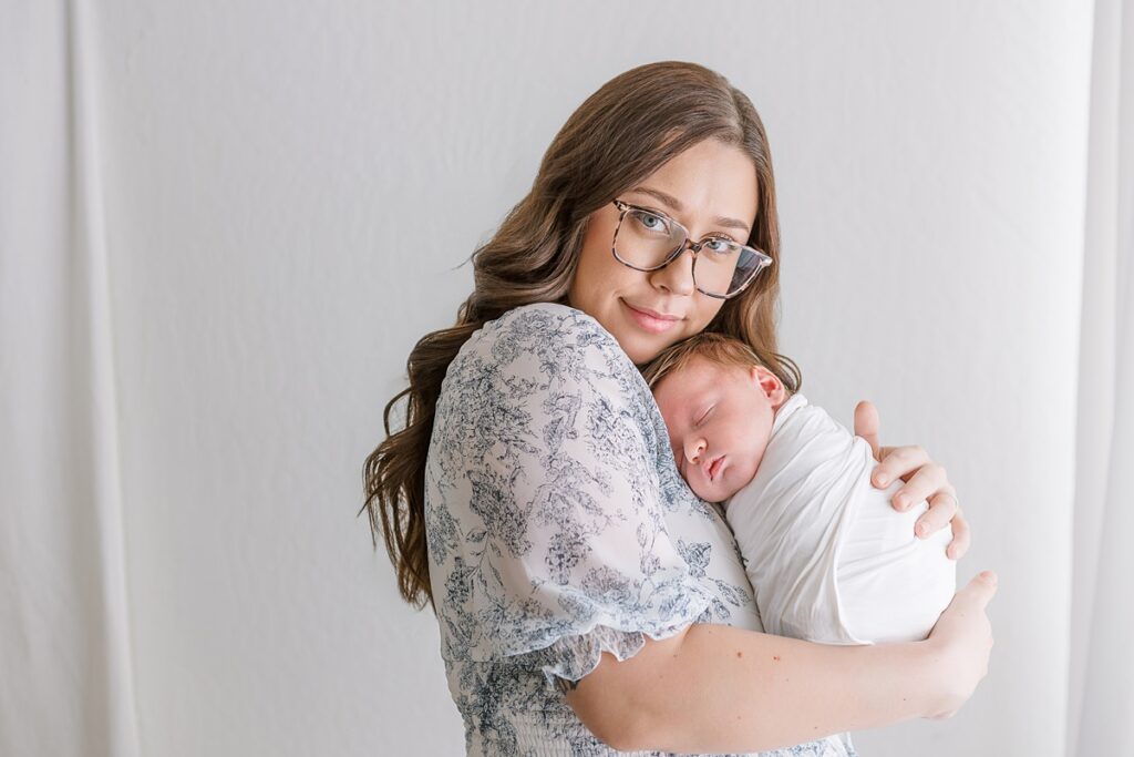 Mother cuddling her newborn baby boy in a bright white studio, holding him close with a tender expression.