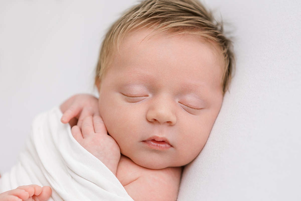 Close-up of newborn baby boy’s face, lying on a soft white blanket in studio light.