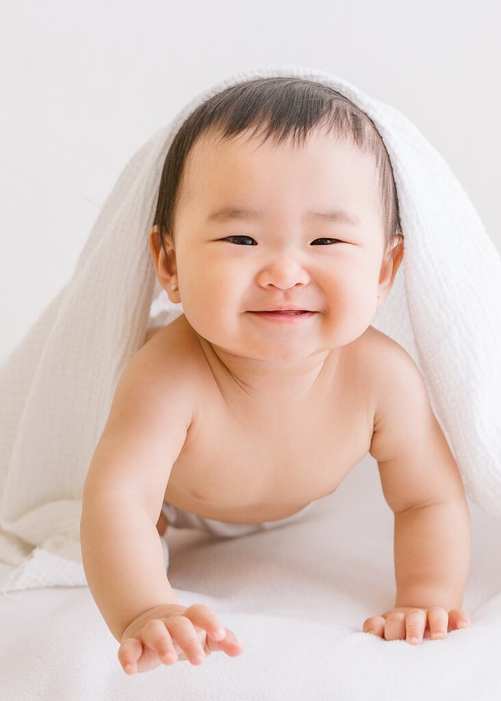 Playful baby girl with soft blanket draped over her head during milestone session