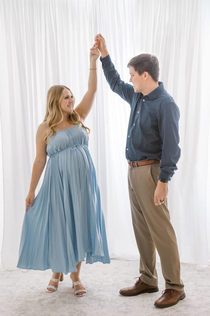 Expecting couple twirling playfully, mom in a blue maternity gown and dad in a blue shirt, photographed in a light-filled white studio.